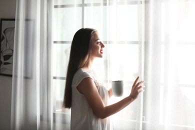 Photo of Young woman standing near window with curtains at home