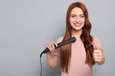 Beautiful woman with hair iron showing thumbs up on light gray background, space for text