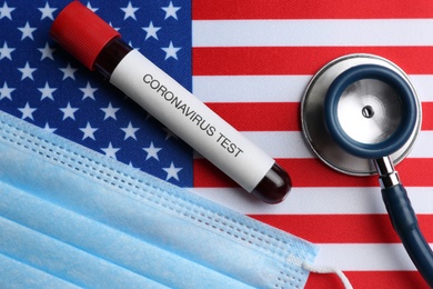 Photo of Test tube with blood sample, medical mask and stethoscope on American flag, flat lay. Coronavirus pandemic in USA