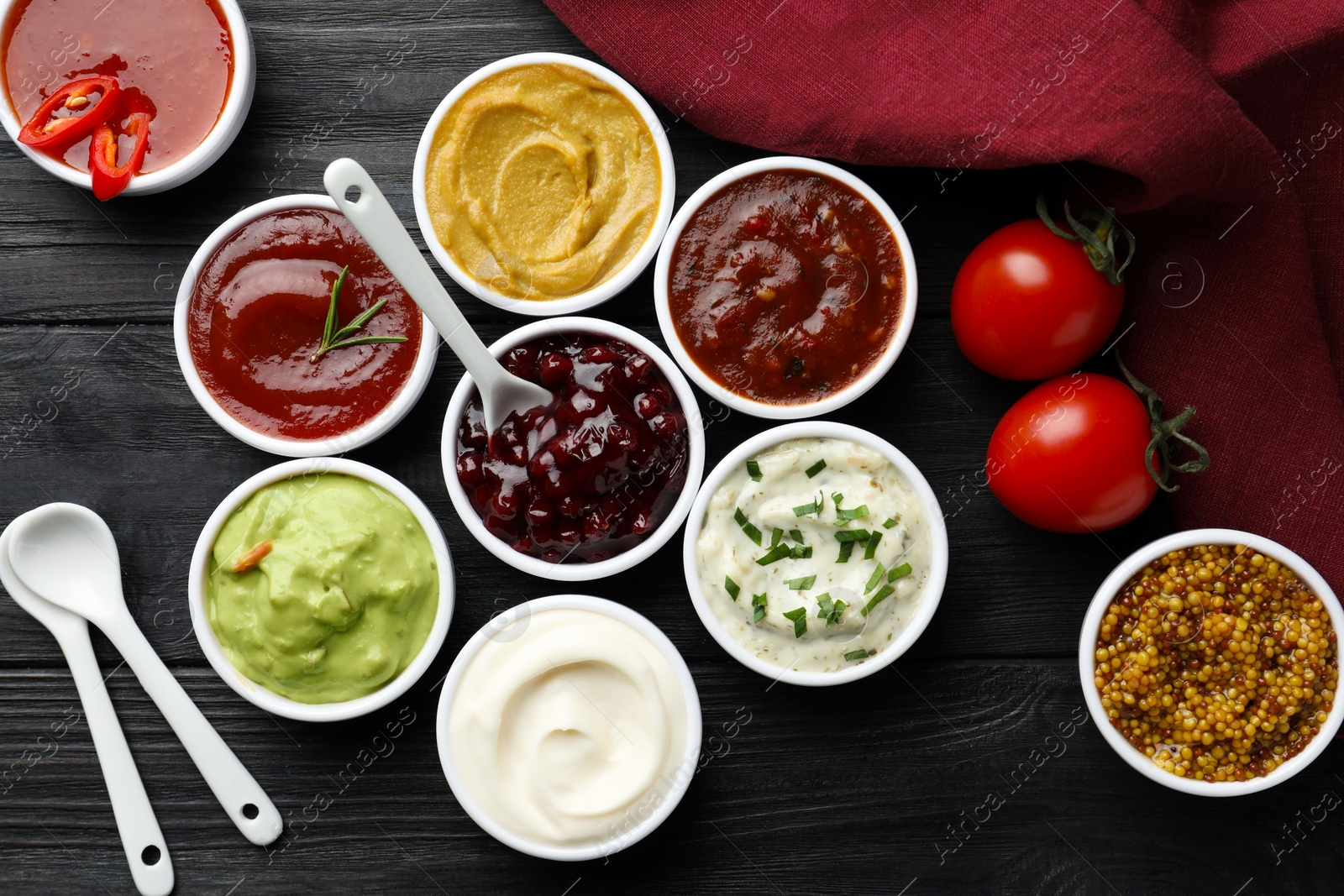 Photo of Different tasty sauces in bowls, tomatoes and spoons on black wooden table, flat lay