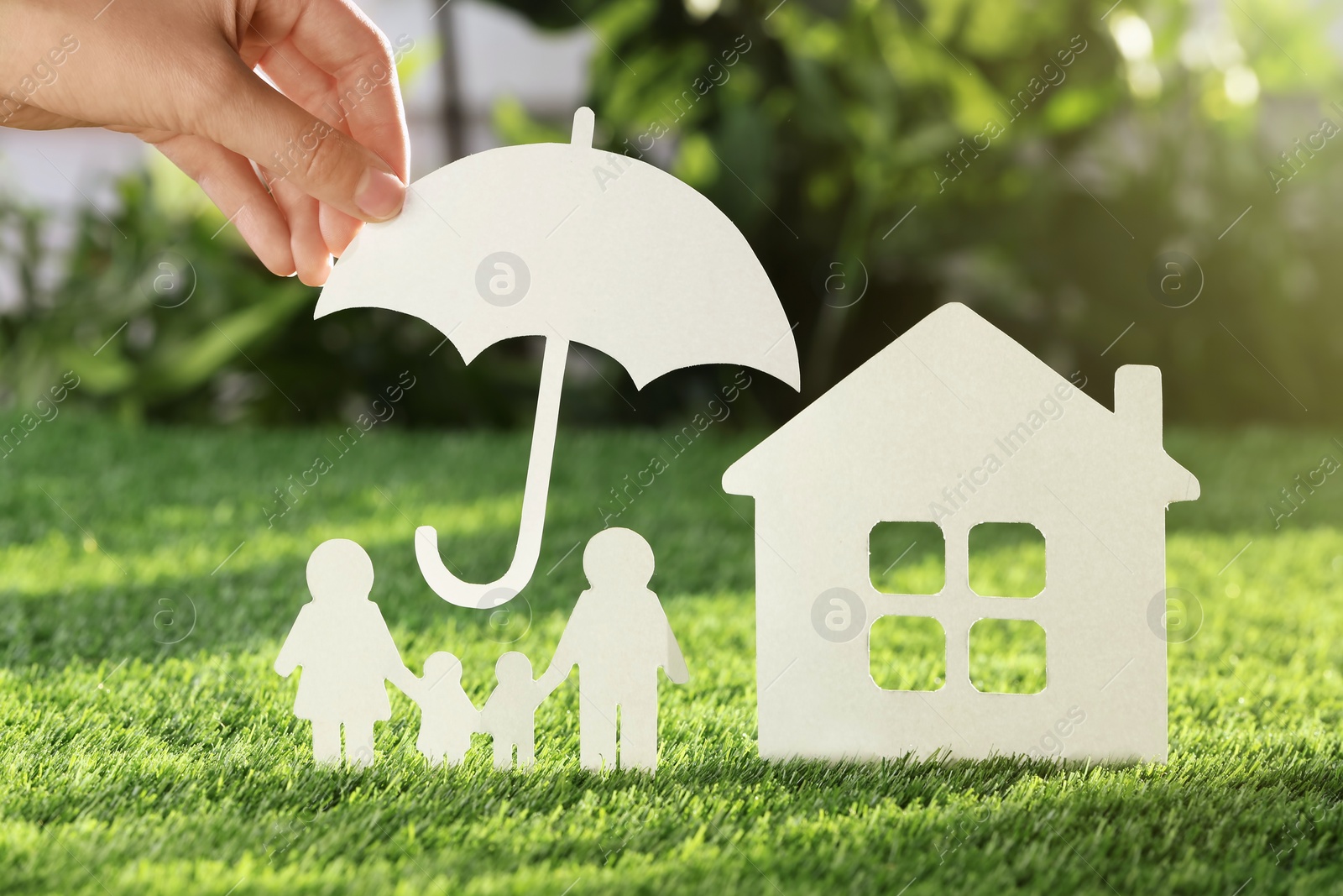 Photo of Woman holding paper umbrella over cutout of family and house on fresh grass, closeup. Life insurance concept