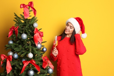 Photo of Beautiful young woman in Santa hat with glass of champagne near Christmas tree on color background