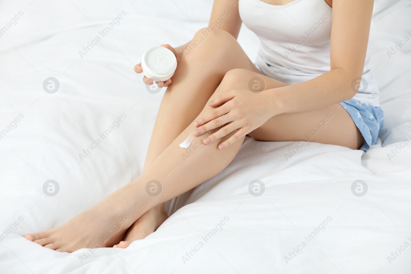 Photo of Young woman applying body cream onto leg on bed, closeup