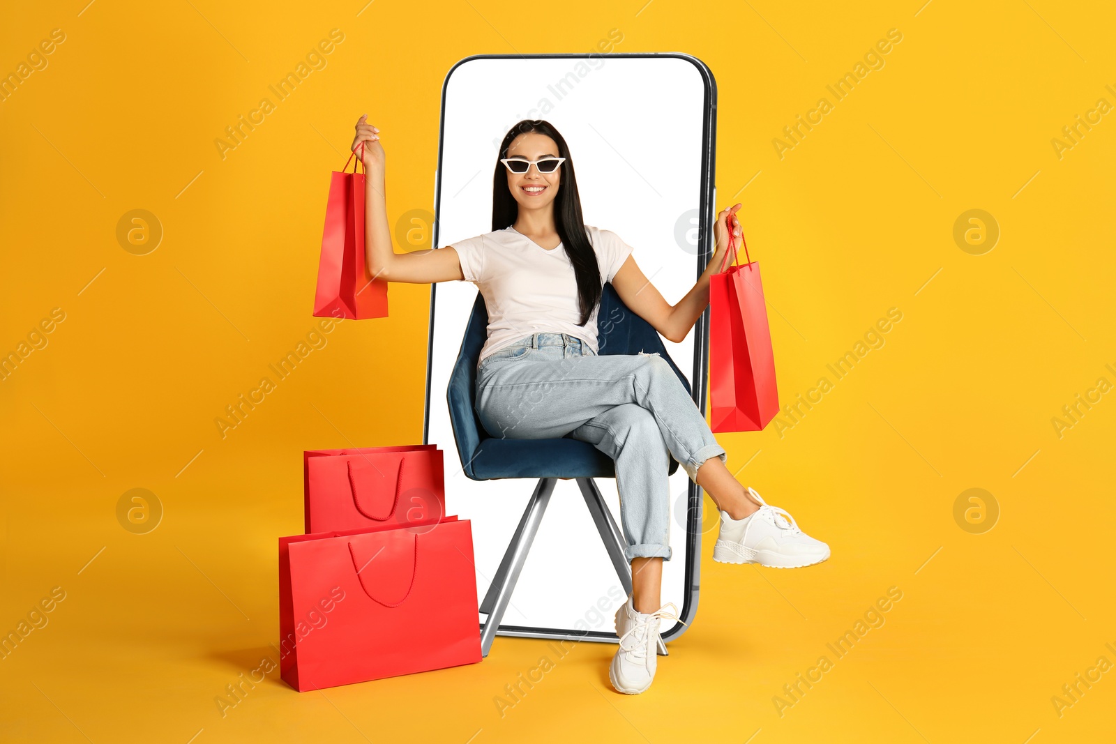 Image of Online shopping. Happy woman with paper bags sitting in armchair near smartphone on orange background