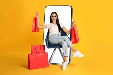 Image of Online shopping. Happy woman with paper bags sitting in armchair near smartphone on orange background