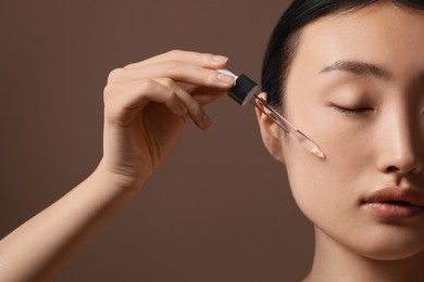 Beautiful young woman applying cosmetic serum onto her face on brown background, closeup