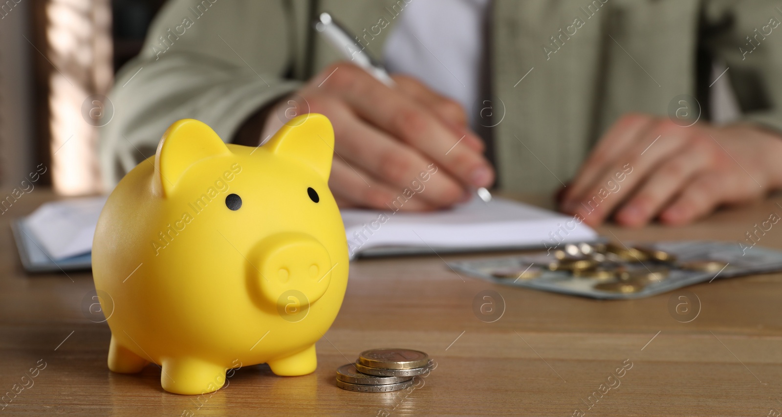 Photo of Man at wooden table, focus on yellow piggy bank. Space for text