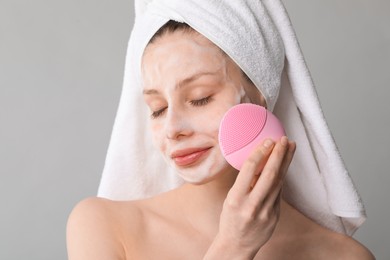 Photo of Young woman washing face with brush and cleansing foam on grey background