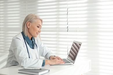Photo of Mature female doctor working with laptop at table in office