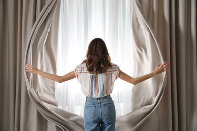 Woman opening window curtains at home in morning, back view