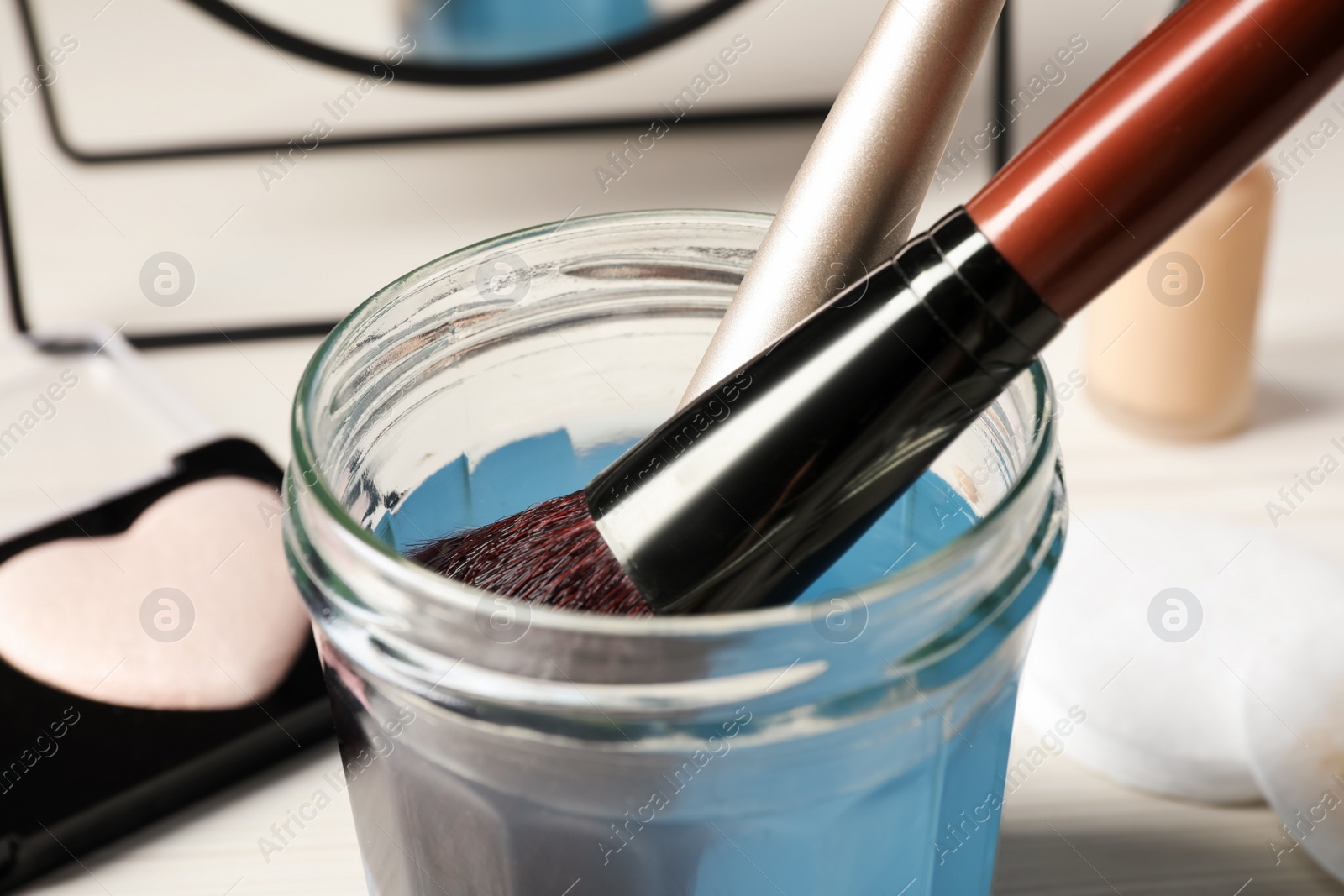 Photo of Cleaning makeup brushes in jar with special liquid on white wooden table, closeup