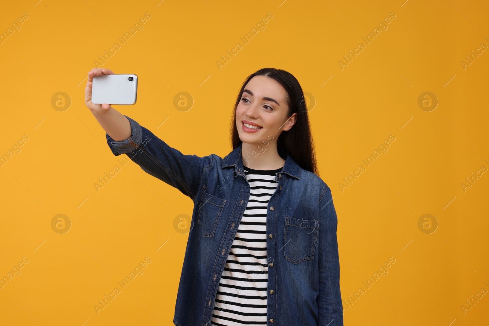 Photo of Smiling young woman taking selfie with smartphone on yellow background