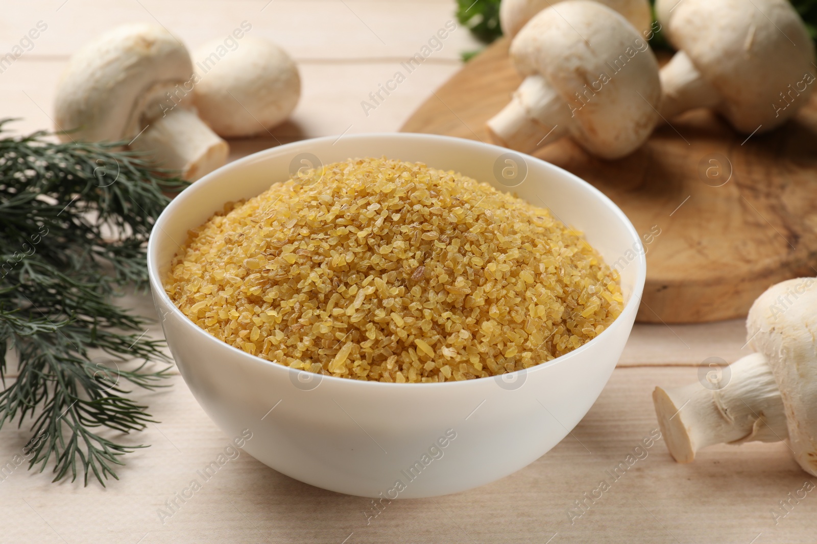 Photo of Raw bulgur in bowl, mushrooms and dill on light wooden table, closeup