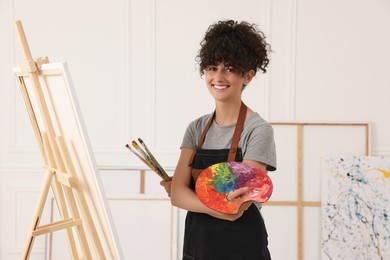 Young woman holding brushes and artist`s palette near easel with canvas