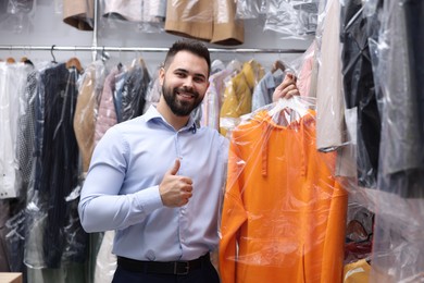 Dry-cleaning service. Happy worker holding hanger with hoodie in plastic bag and showing thumb up near other clothes indoors