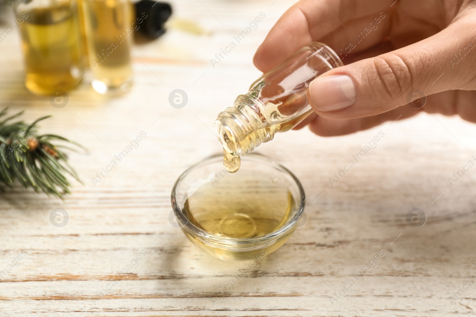 Photo of Woman pouring conifer essential oil into bowl on white wooden table, closeup