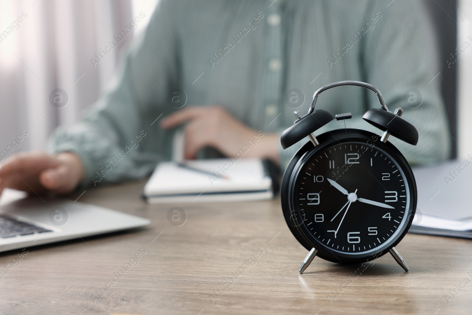 Photo of Black alarm clock and woman working on laptop at table, closeup. Space for text