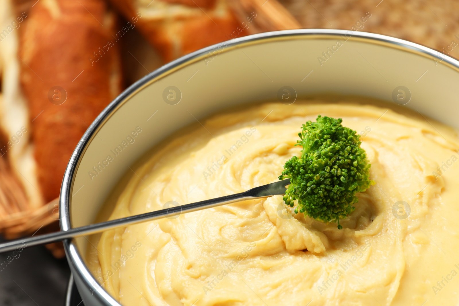 Photo of Dipping piece of broccoli into fondue pot with melted cheese on blurred background, closeup