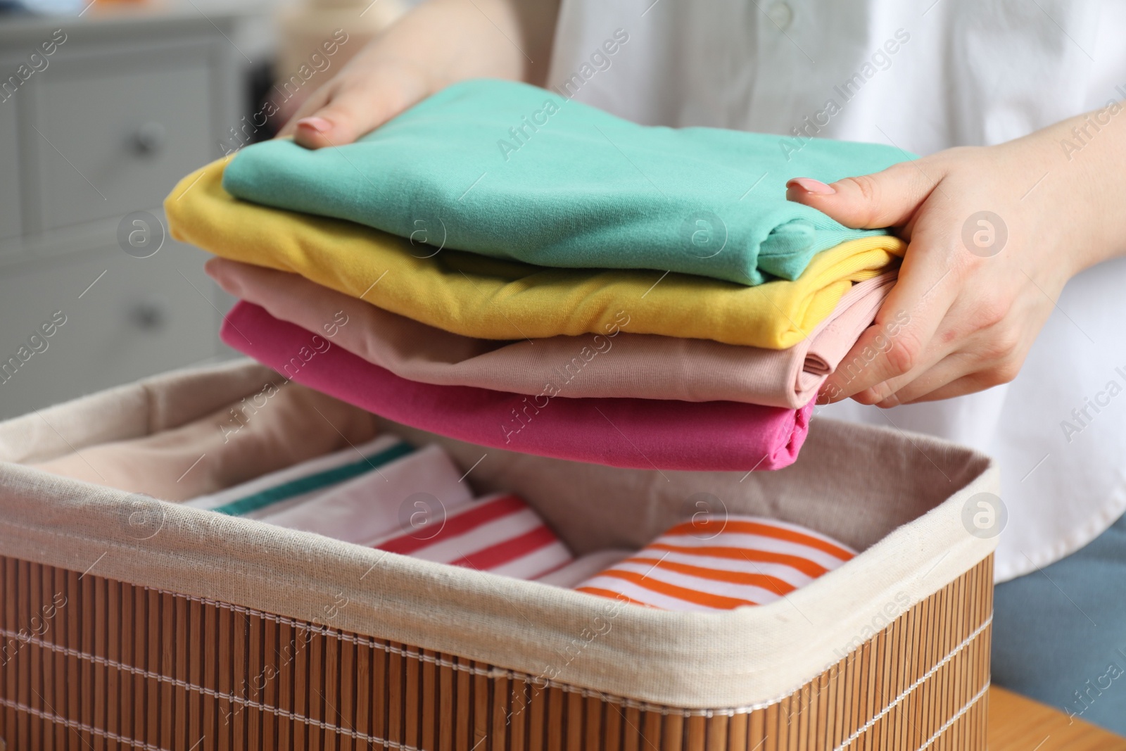 Photo of Woman putting folded clothes into storage basket at table indoors, closeup