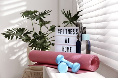Photo of Sport equipment and lightbox with hashtag FITNESS AT HOME on window sill indoors. Message to promote self-isolation during COVID‑19 pandemic