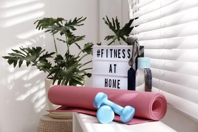 Sport equipment and lightbox with hashtag FITNESS AT HOME on window sill indoors. Message to promote self-isolation during COVID‑19 pandemic