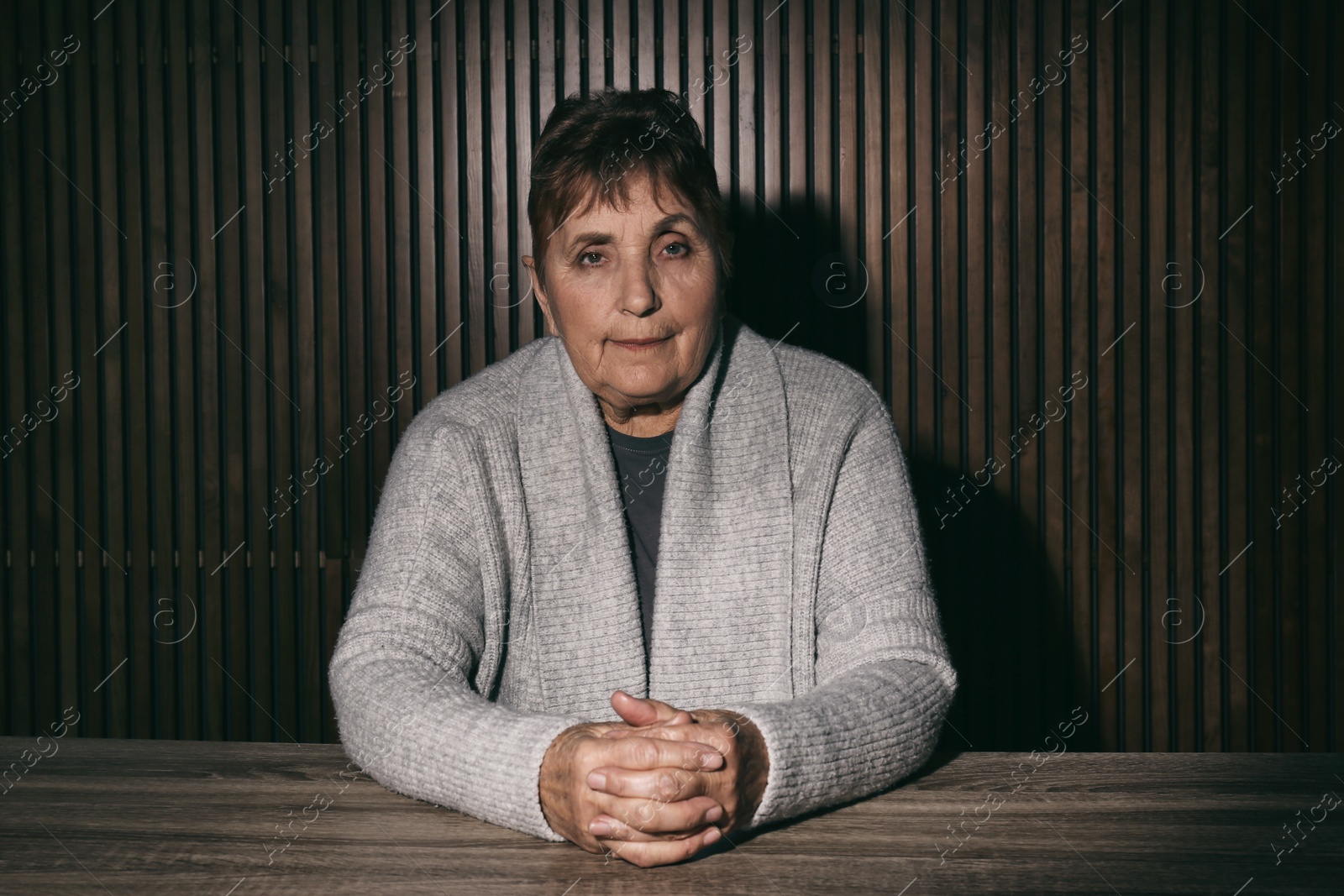 Photo of Portrait of poor senior woman sitting at table against wooden wall