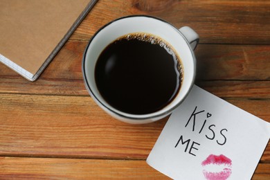 Photo of Sticky note with phrase Kiss Me, lipstick mark and cup of coffee on wooden table, above view