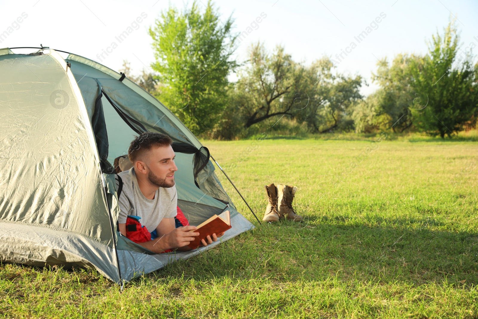 Photo of Young man in sleeping bag with book lying inside camping tent