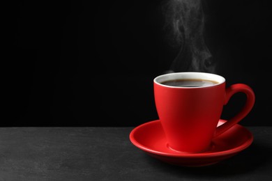 Red cup with hot steaming coffee on grey table against black background, space for text