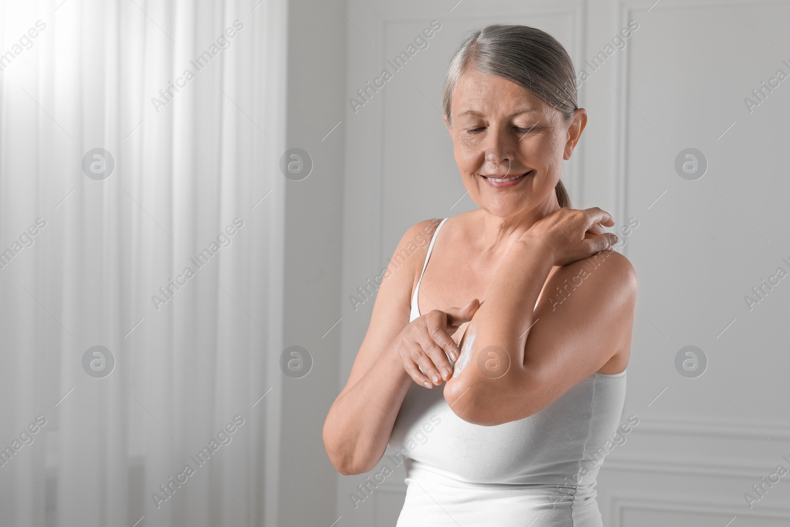 Photo of Happy woman applying body cream on elbow indoors. Space for text