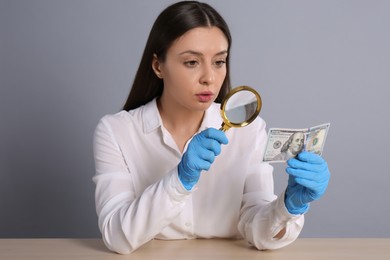 Photo of Expert authenticating 100 dollar banknote with magnifying glass at table on light grey background. Fake money concept