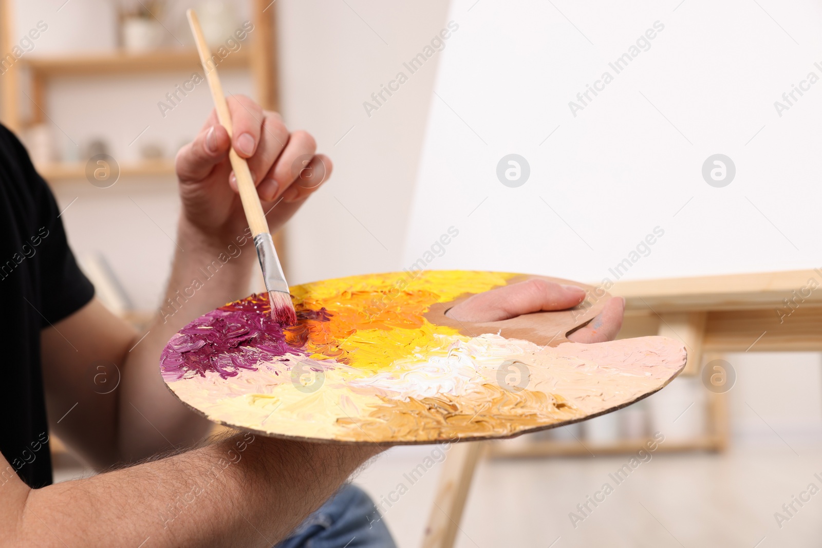 Photo of Man with brush and artist`s palette painting in studio, closeup. Using easel to hold canvas