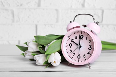 Pink alarm clock and beautiful tulips on white wooden table against brick wall. Spring time