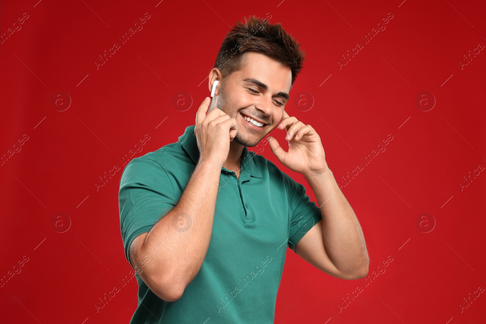 Photo of Happy young man listening to music through wireless earphones on red background