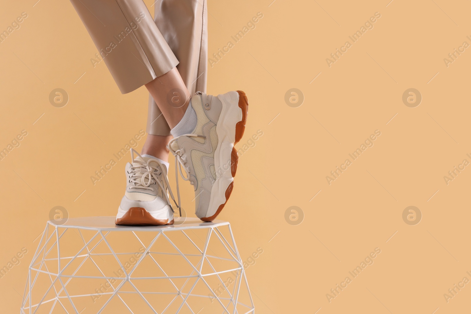 Photo of Woman in stylish new sneakers standing on white table against beige background, closeup. Space for text