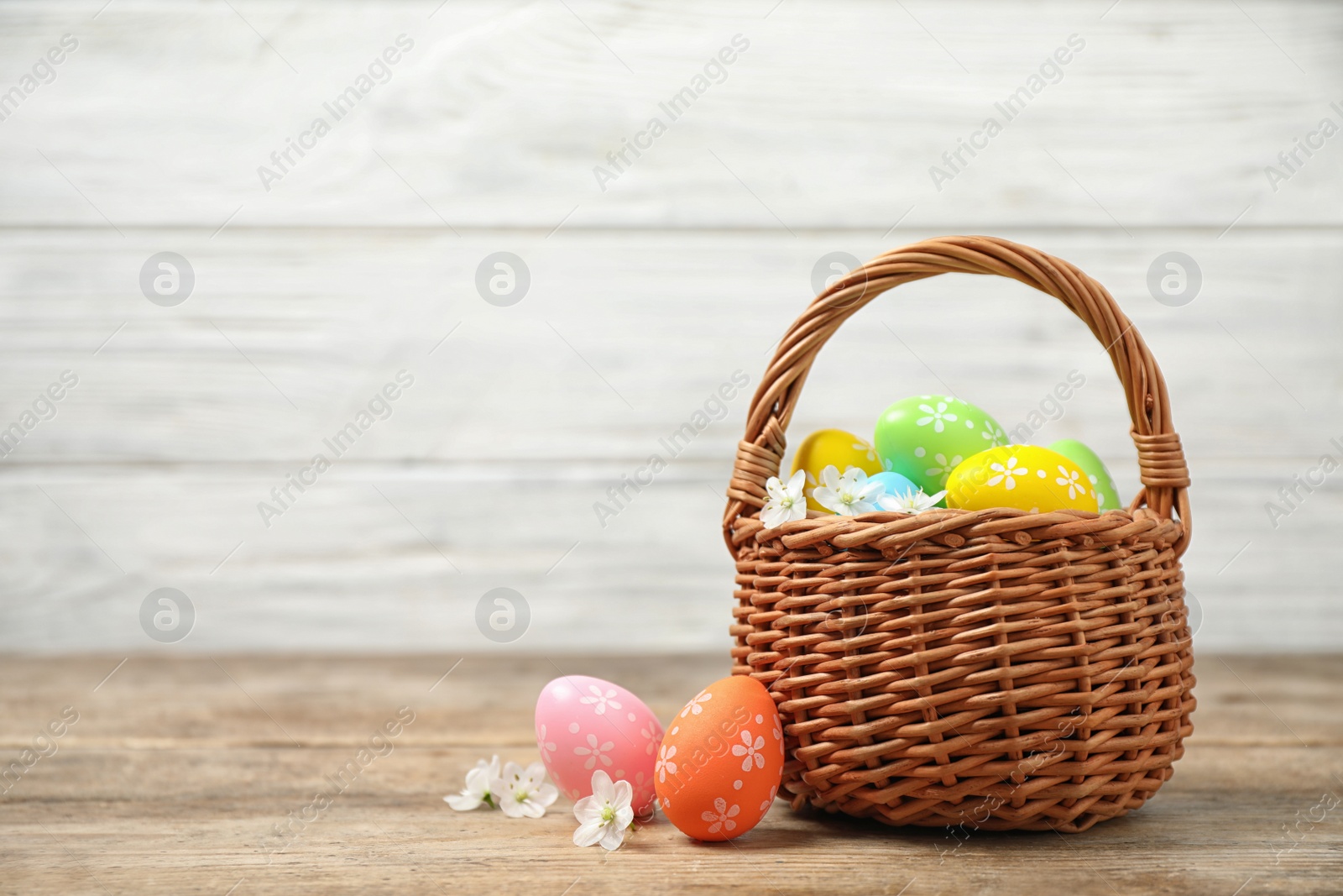 Photo of Colorful Easter eggs in basket and flowers on wooden table. Space for text