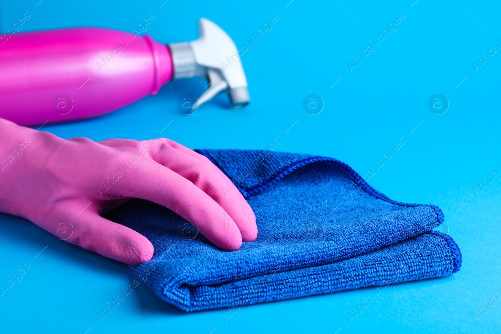 Photo of Woman in pink rubber gloves with microfiber cloth and detergent on light blue background, closeup