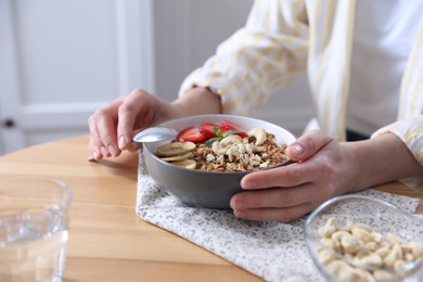 Woman eating tasty granola with banana, cashew and strawberries at wooden table indoors, closeup