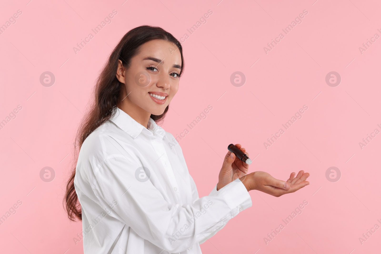 Photo of Beautiful happy woman with roller bottle applying essential oil onto wrist on pink background