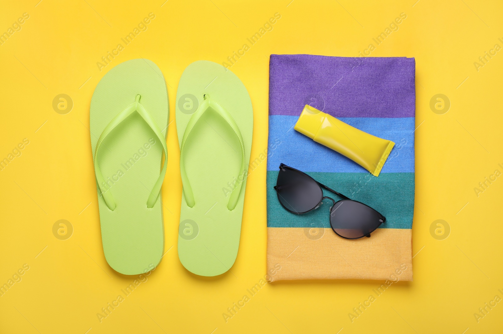 Photo of Flip flops, sunscreen, stylish sunglasses and striped towel on yellow background, flat lay