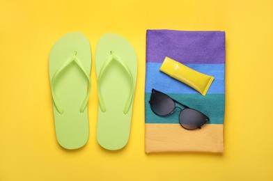 Photo of Flip flops, sunscreen, stylish sunglasses and striped towel on yellow background, flat lay