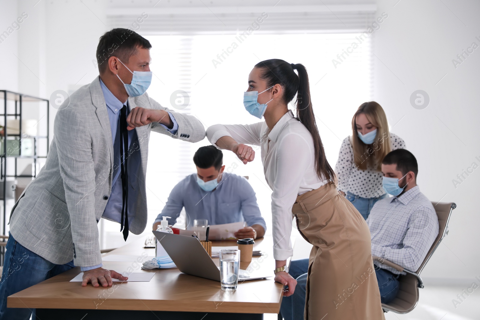 Photo of Coworkers with protective masks making elbow bump in office. Informal greeting during COVID-19 pandemic