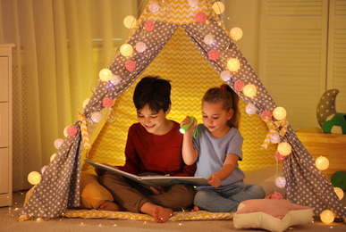 Photo of Little children with flashlight reading book in play tent at home