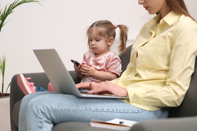 Photo of Mother working remotely on laptop while her daughter playing with smartphone at home, closeup