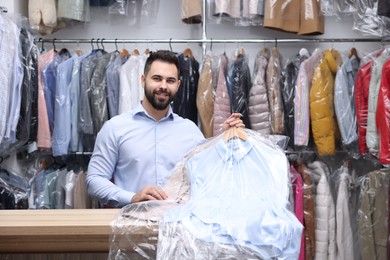 Photo of Dry-cleaning service. Happy worker holding hangers with clothes in plastic bags at counter indoors