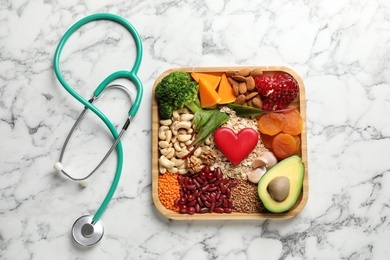Photo of Wooden tray with heart-healthy products and stethoscope on marble background, flat lay
