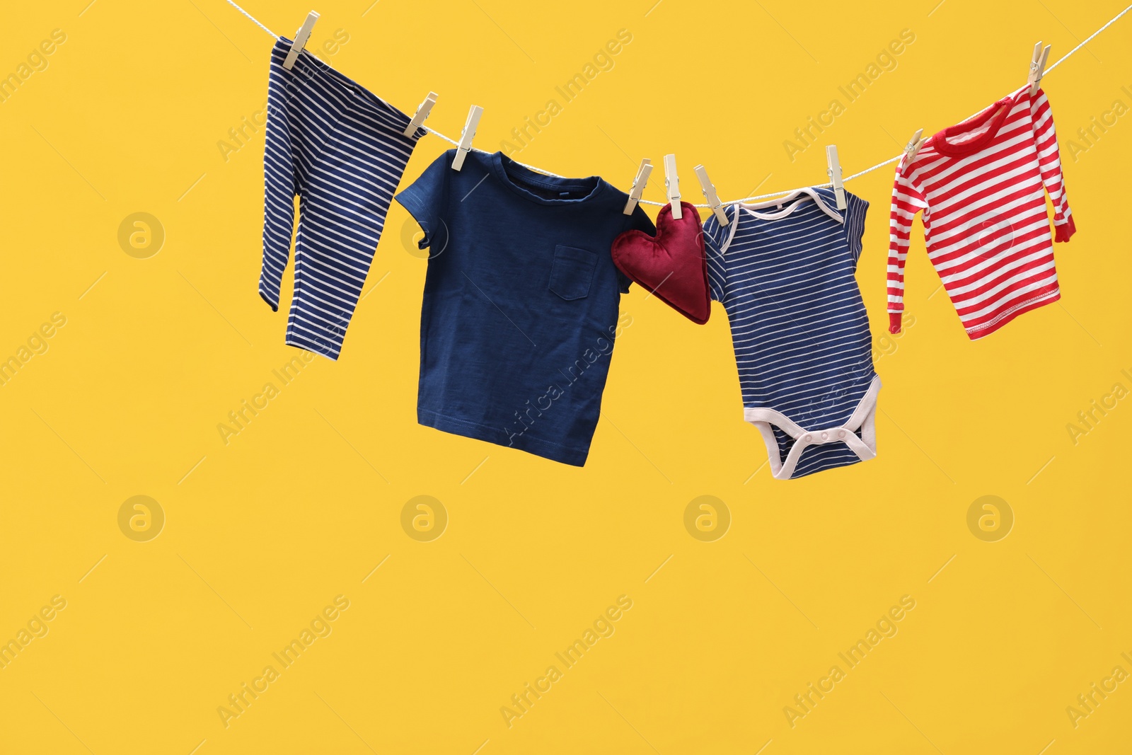 Photo of Different baby clothes and heart shaped cushion drying on laundry line against orange background