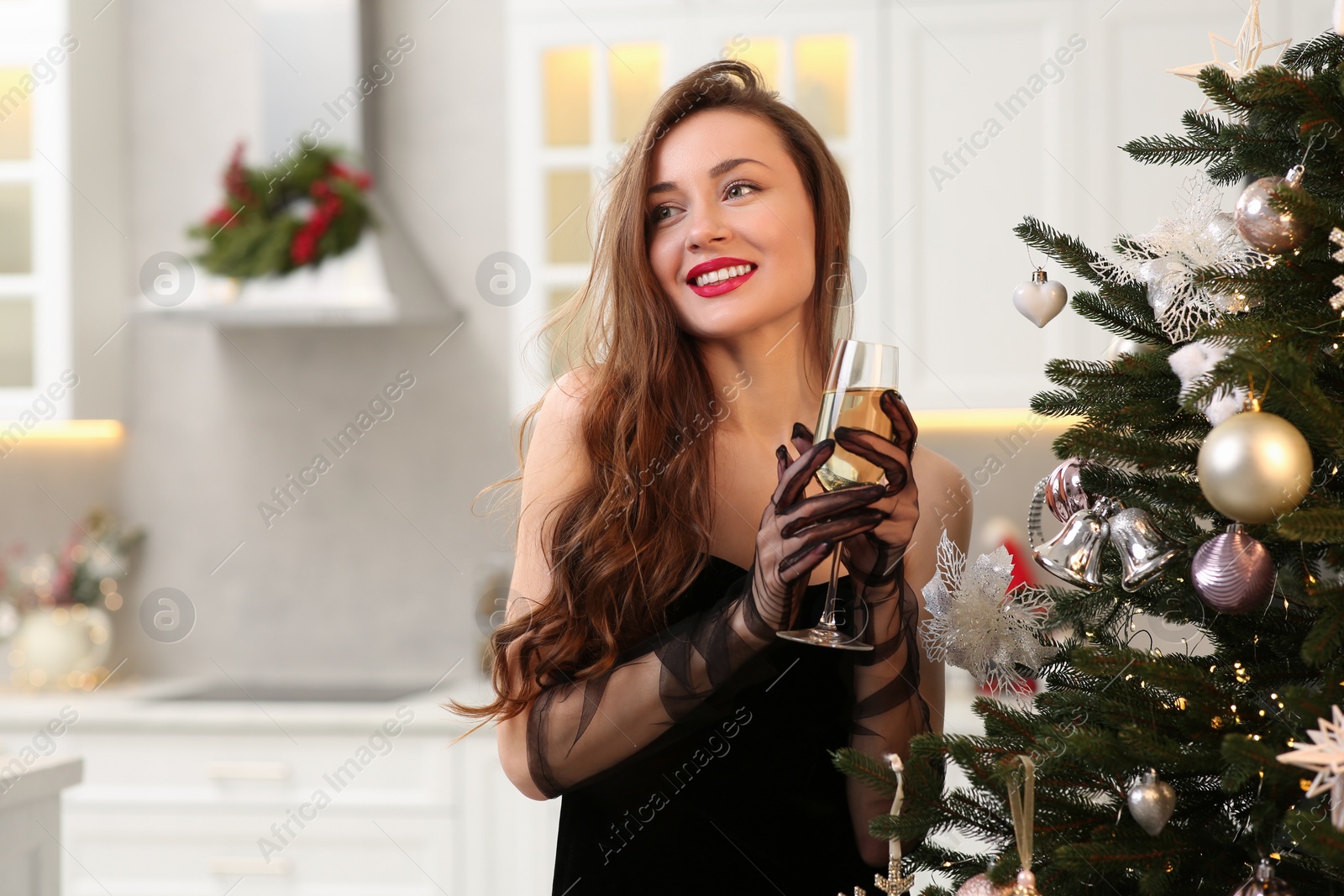 Photo of Beautiful young smiling woman with glass of sparkling wine near Christmas tree at home, space for text