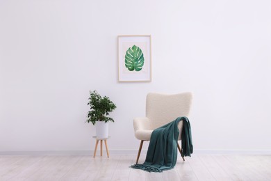 Photo of Comfortable armchair, blanket, houseplant and picture on white wall indoors