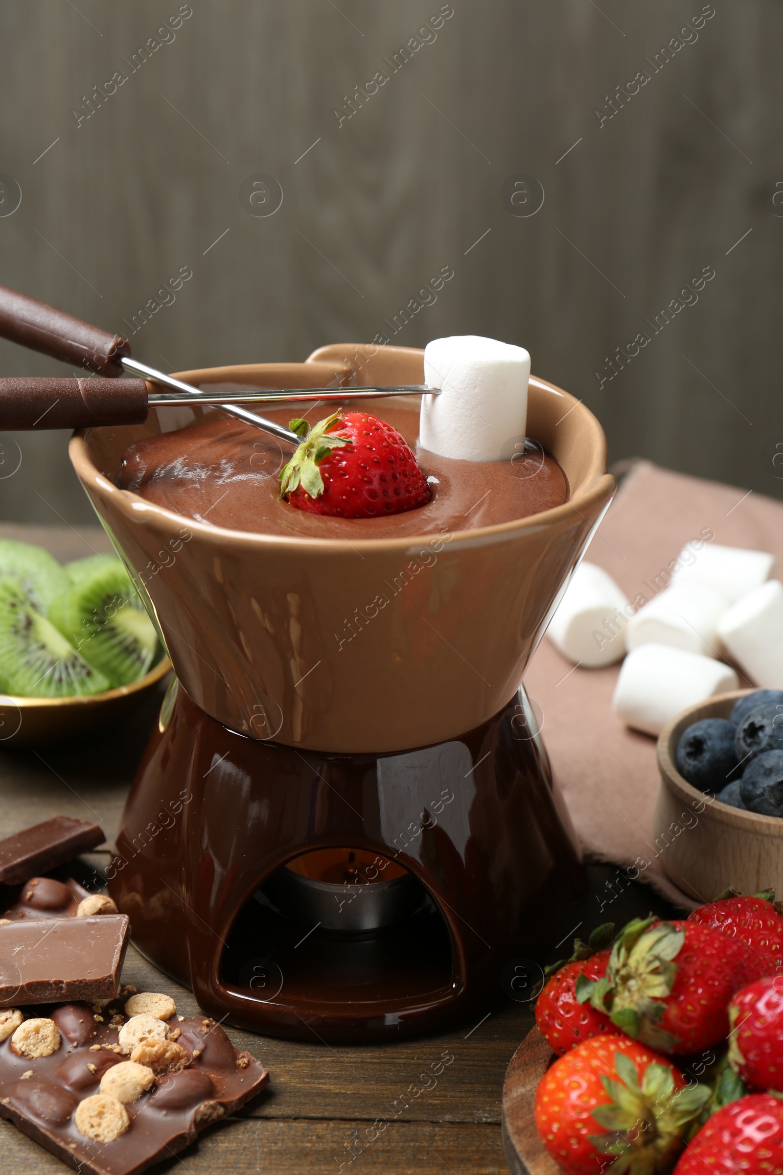 Photo of Fondue pot with melted chocolate, different fresh berries, kiwi, marshmallows and forks on wooden table